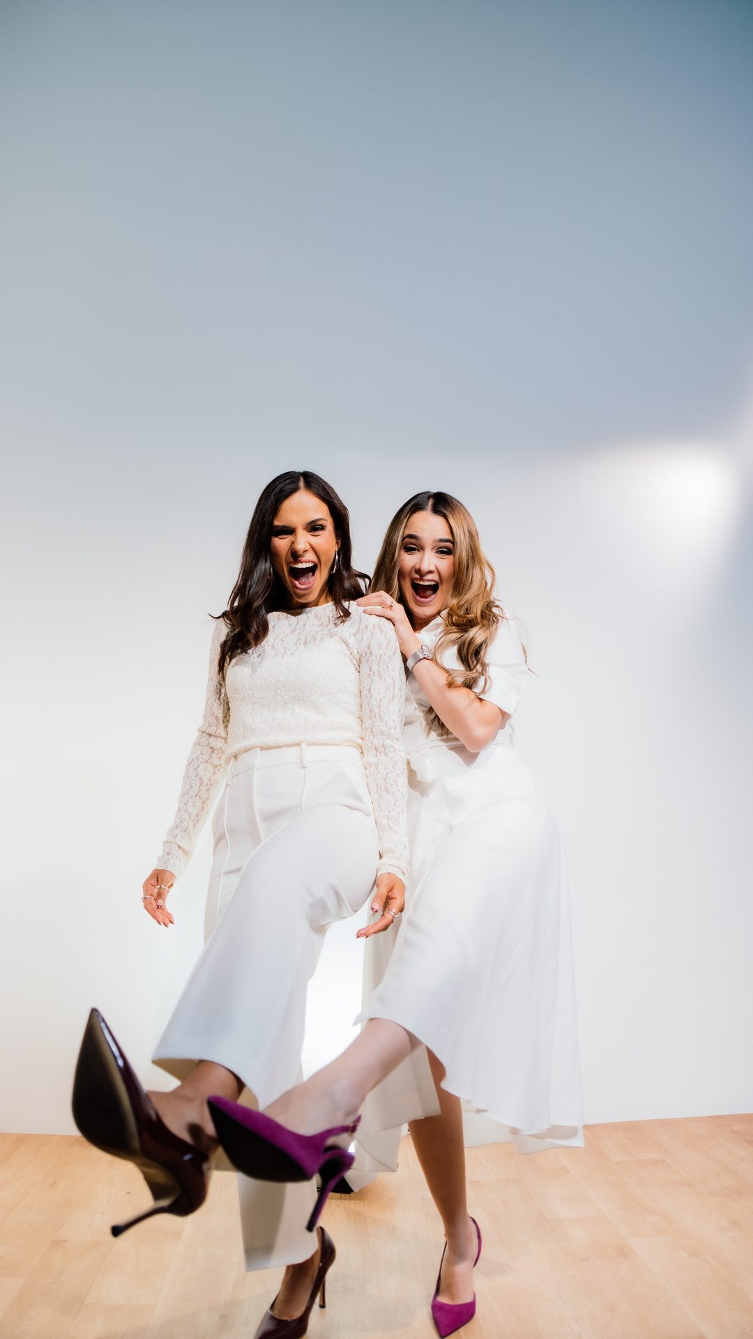 Ana Gabriela Fidalgo y Michelle Ruiz, vestidas de blanco, de cuerpo entero y riendo en un estudio de fondo claro.